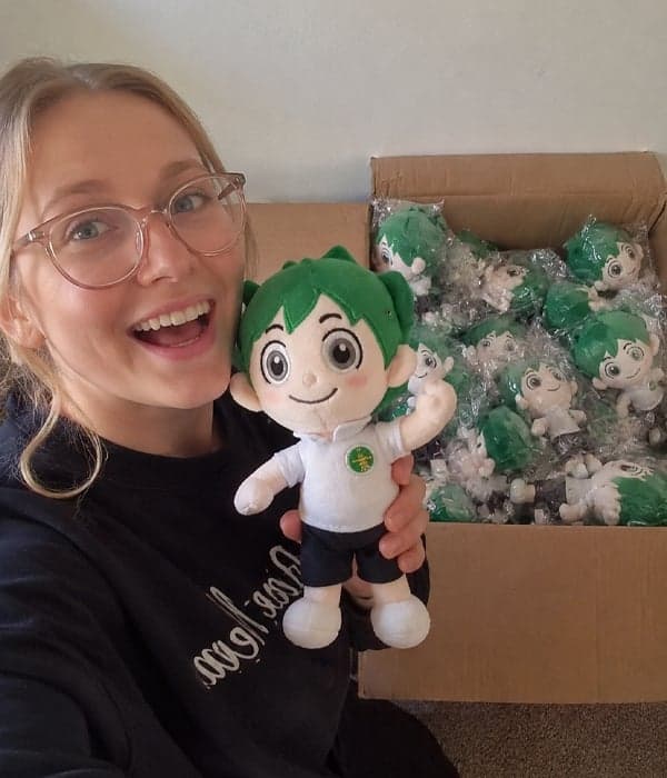 Smiling client unboxing a bulk delivery of custom green-haired anime boy mascot plush toys with embroidered chest logo, manufactured by StarDream Toys